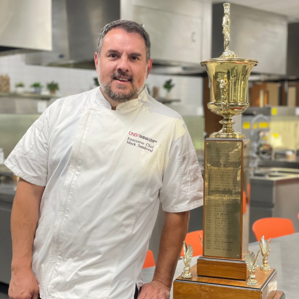 A man (Mark Sandoval) in a chef's jacket stands next to the Chef of the Year Award.