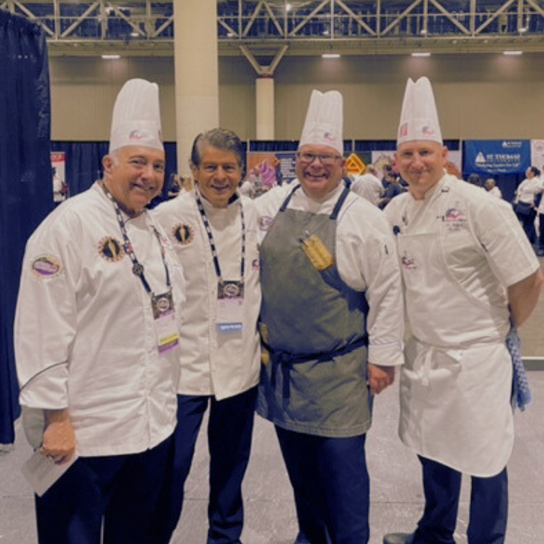 Four make chefs pose standing for a picture with a convention happening in the background.