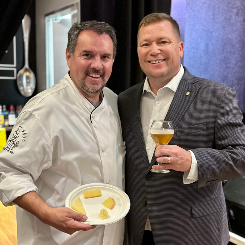 A male chef stands holding a plate of cheese while another male in a suit holds a glass of beer.