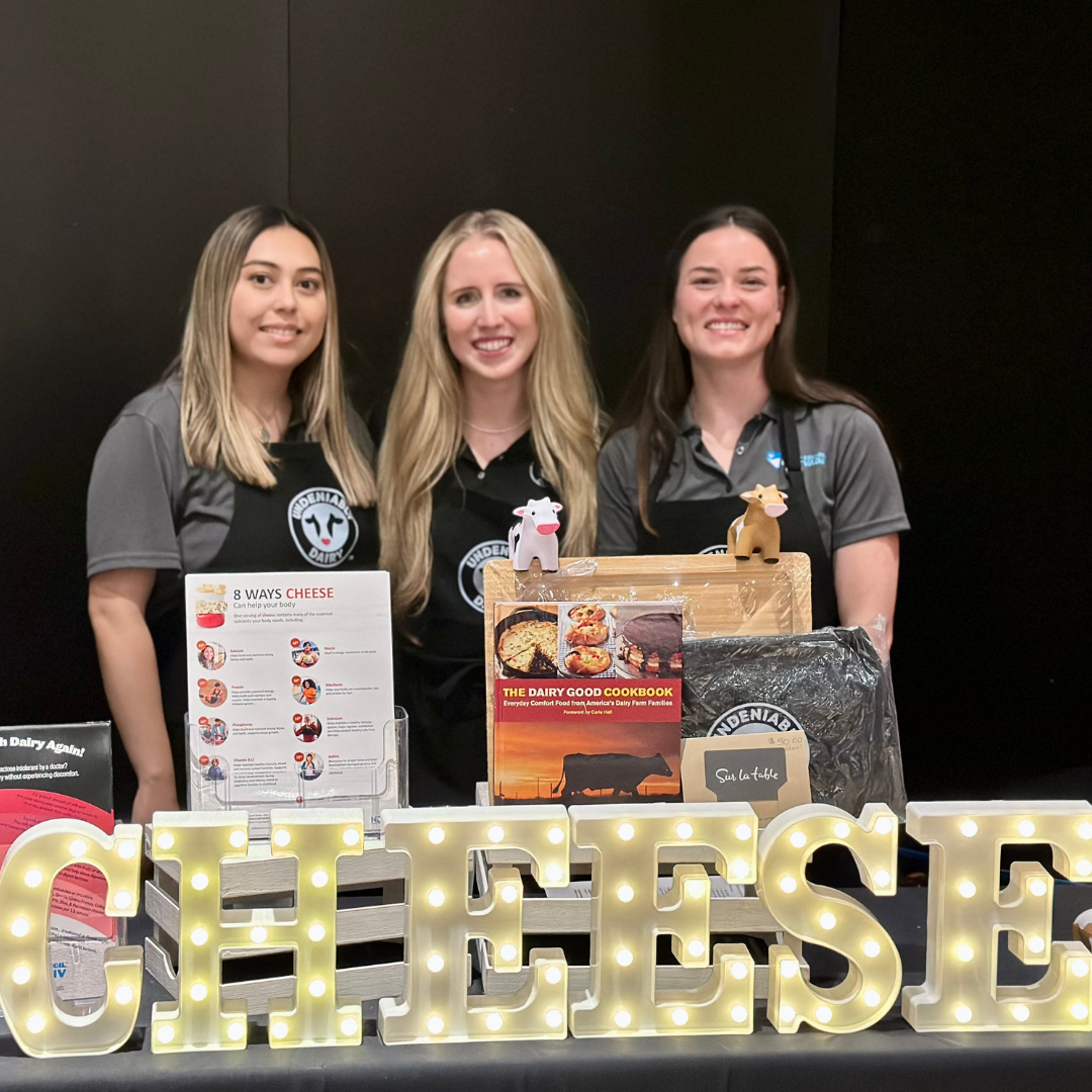 Three women stand behind a booth including a lit sign that says "Cheese".