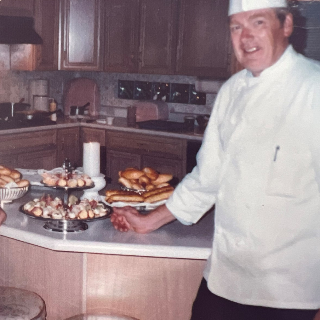 A man in a chef coat and hat poses by a table full of pastries.